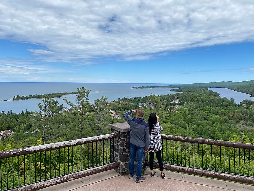A couple peers out from a roadside lookout over a small, heavily forested town on the shore of Lake Superior.