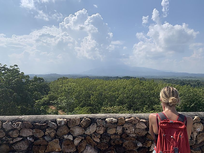 A blond female backpacker looks out over the jungle from atop a stone tower