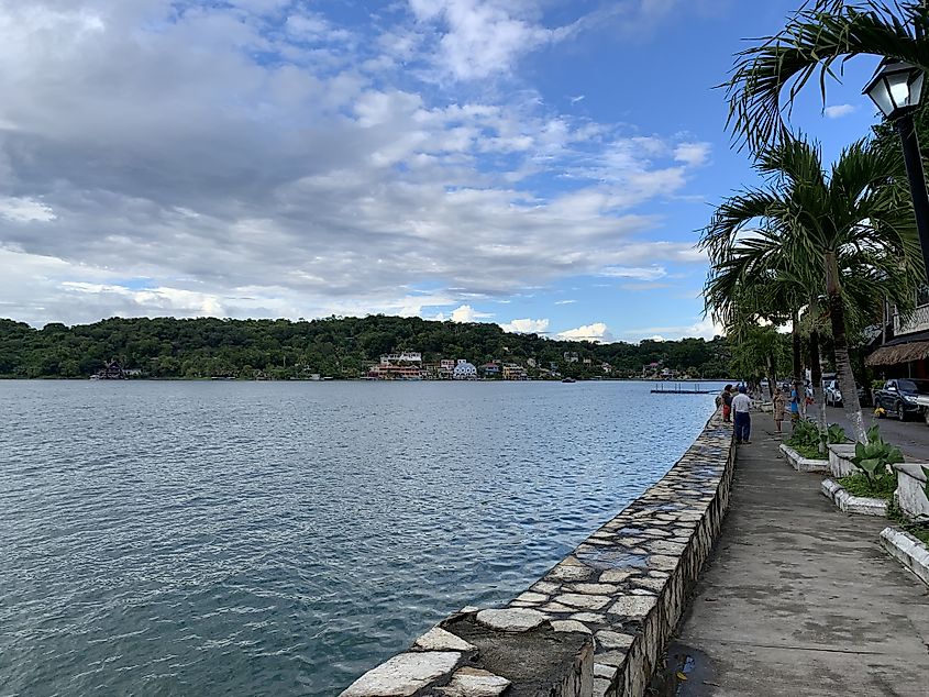 A waterside walkway with small, colorful homes across the bay and casual pedestrians in front.