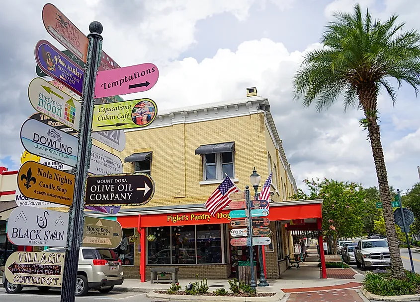 Colorful directional signpost with business names in downtown Mount Dora, Florida, in front of a yellow brick building with red awnings and American flags.