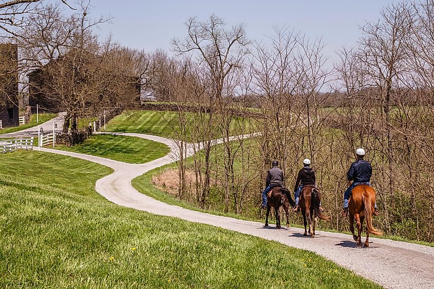 Three adult riders guide their horses along a trail winding across farmland at the landmark destination of Shaker Village of Pleasant Hill