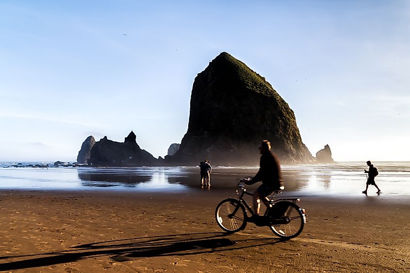 Haystack Rock at Cannon Beach on the Oregon Coast, with a silhouetted couple taking a selfie and a bicyclist passing by in blurred motion