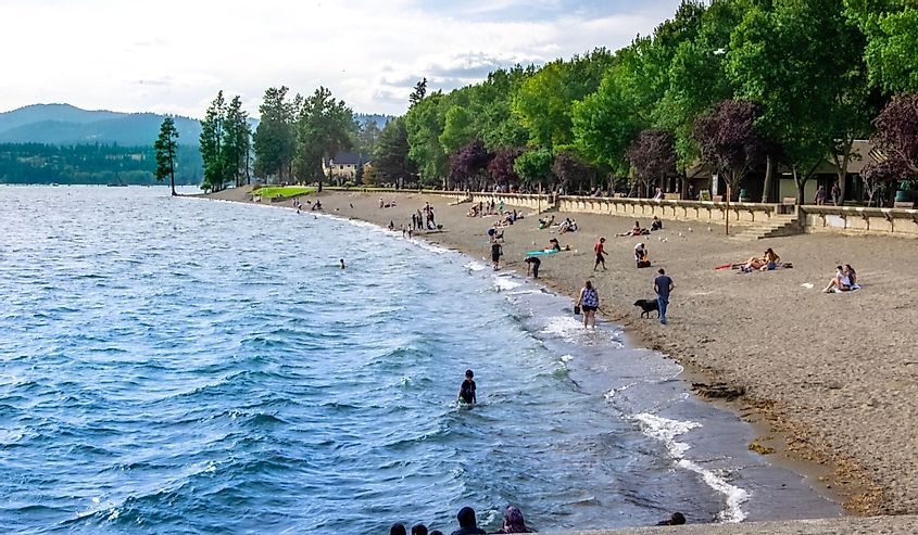 In Lake Coeur D'Alene visitors relax on the beach and play in the water at public beach along alpine lake in northern Idaho with trees and mountains in the background.