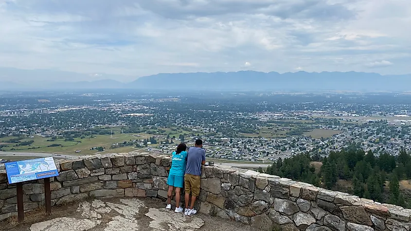 A couple leans against a rock wall, looking out from the bluff over an adjacent mountain range and mountain town below.