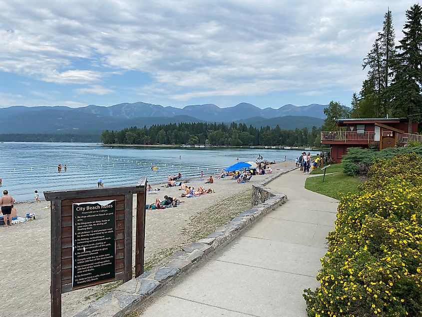 A small lake beach backdropped by mountains and lined with wild flowers draws a crowd on a pleasant, overcast day.  