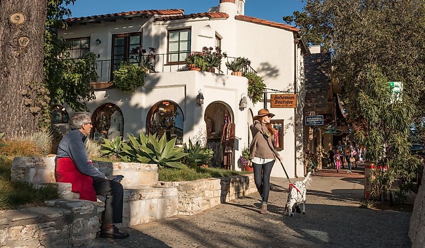 A fashionably-dressed, tall and slender woman strolls down a sidewalk and a senior gentleman sitting on a bench in Carmel on a November afternoon.