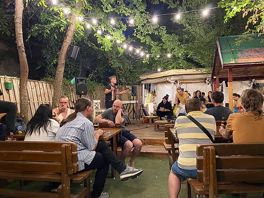 A poet stands on a small stage, reading to an audience in an outdoor social space.