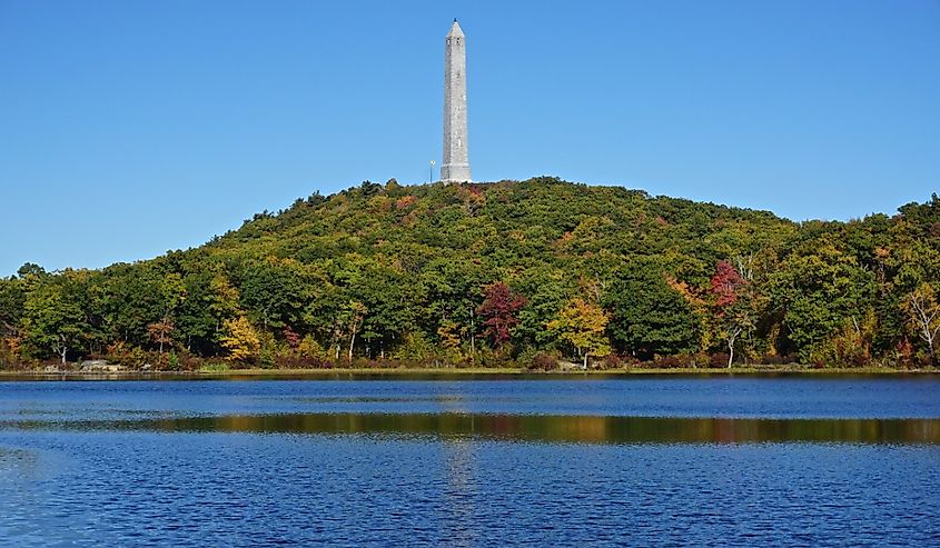 An obelisk-shaped veterans monument overlooks Lake Marcia, surrounded by fall foliage at High Point State Park, Montague.
