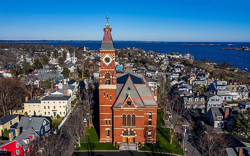 Aerial view of a coastal town featuring a prominent red-brick church with a tall clock tower. Surrounding it are quaint houses, with the ocean in the backdrop. Bright, clear sky adds a vibrant, serene feel.