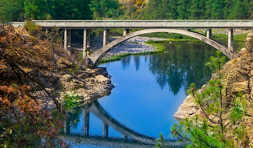 Arched concrete bridge spans the Spokane river with blue sky and architecture reflecting off of the water in time exposure with evergreen trees in background.