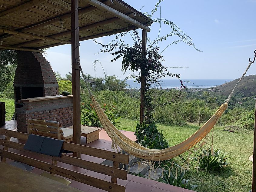 A hilltop patio area, with a picnic table, a wood burning bbq and a yellow hammock overlooking the ocean below. 