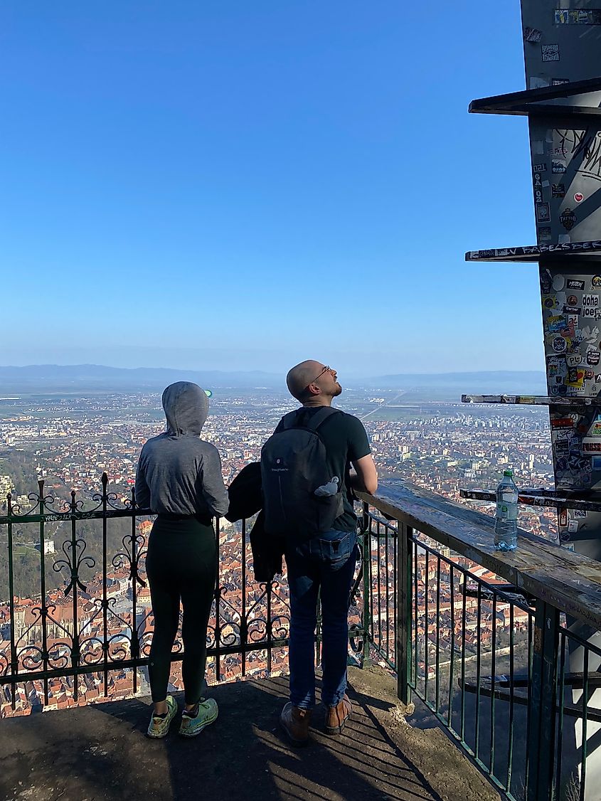 Two hikers stand atop a mountain observation platform, looking out over an orange-roofed city.