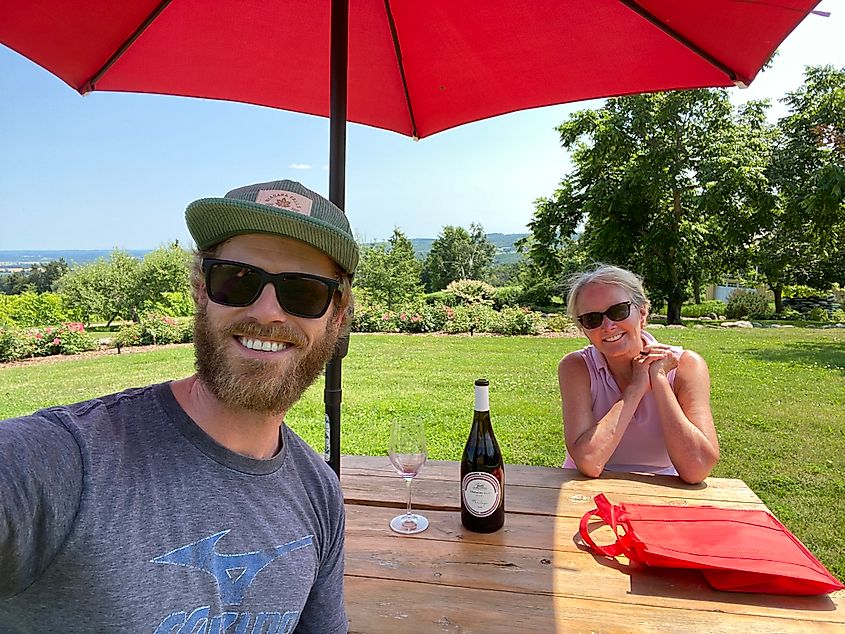 A man takes a selfie with a woman at a picnic table with a bottle of red wine on it.