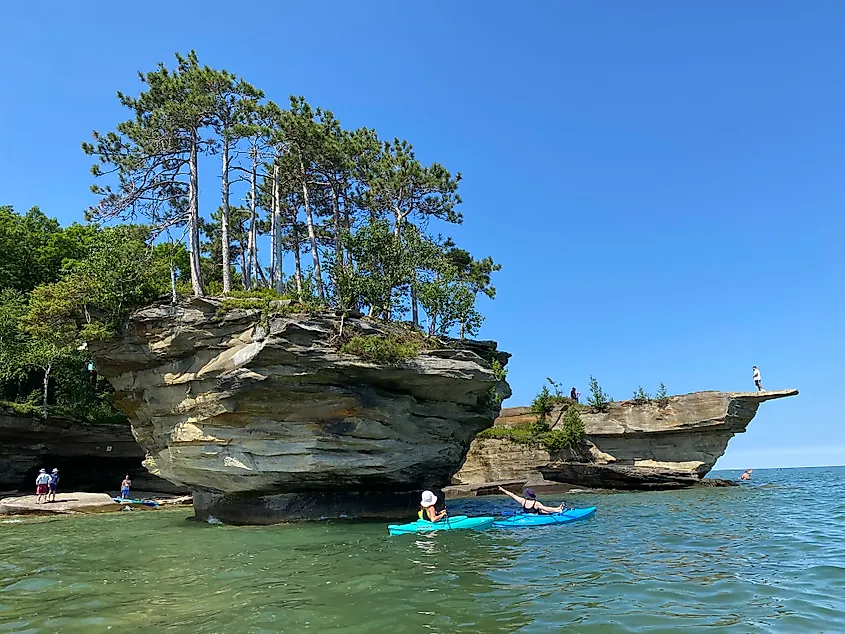 Kayakers circle a floating, striated island while other people investigate the caves and viewpoints on the shore. 