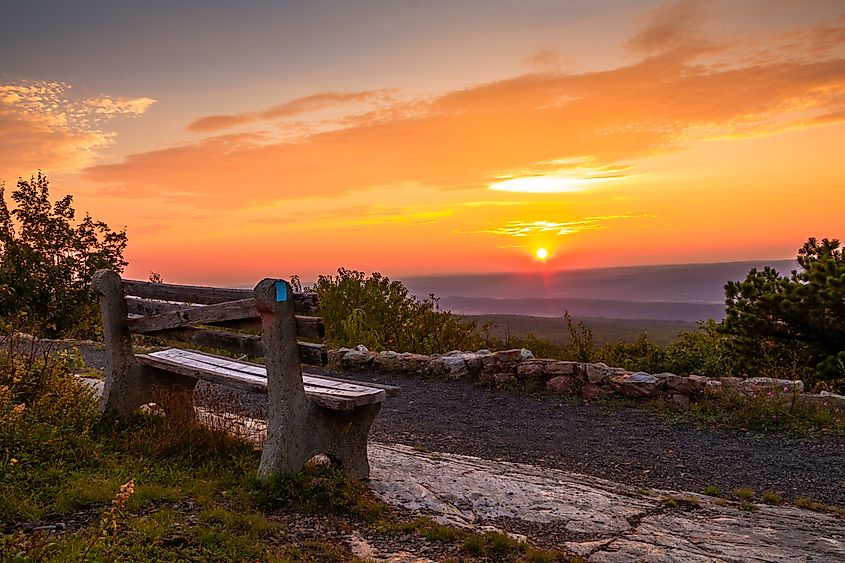 A stone bench overlooks a stunning sunset with vibrant orange and pink hues. The sky is partially cloudy, casting a peaceful glow over the landscape.