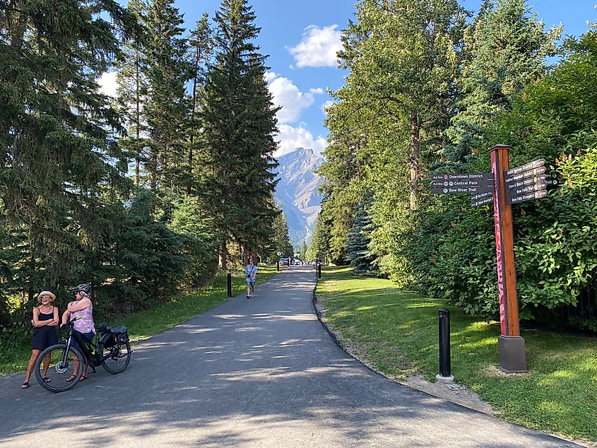 A mountain biker stops to chat with a walker at a signed crossroads on a paved path through a stretch of Rocky Mountain forest. 