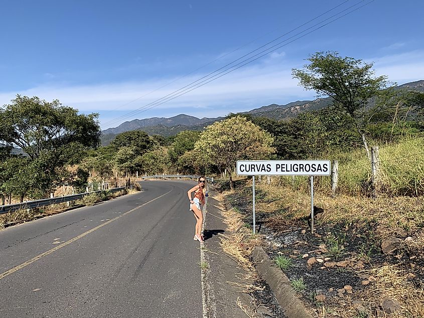 A young woman poses next to a road sign reading "Curvas Peligrosas" in a mountains Mexican countryside