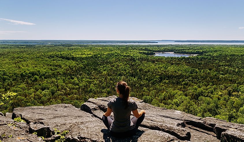 Женщина сидит на вершине скалы и смотрит на лес перед ней в Мичигивадинонг тропа Чашки и блюдца на острове Манитулин, Онтарио, Канада