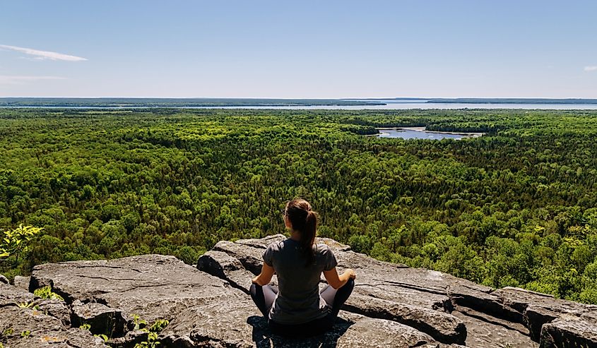 A woman sits on top of a rock and looks at the woods in front of her at Michigiwadinong the Cup and Saucer trail in Manitoulin island, Ontario, Canada