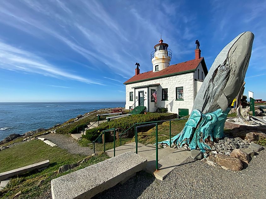 A wooden statue of a breaching whale stands before an old fashioned lighthouse keeper's quarters on a small island before the Pacific Ocean.