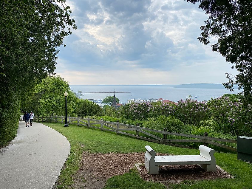 An elderly couple walks up a hill using a paved pedestrian path. At the top, a bench overlooks the village of Mackinac Island and the expansive waters of Lake Huron below.