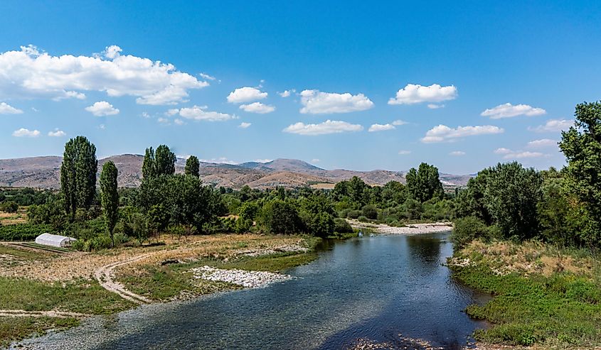  The wide Crna River (Vardar tributary) flows from the distant mountains and bright blue sky beyond. White puffy clouds reflect in the water.