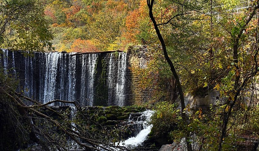 View of the base of Mirror Lake Waterfall. Stone spillway dams lake but allows water to spill over dam. Autumn leaves fill the sky at the Blanchard Springs Caverns Recreation area.