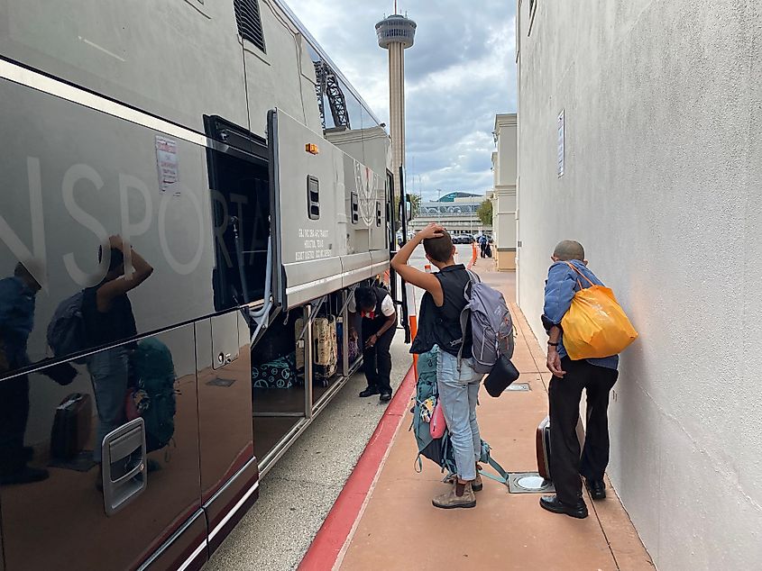 A young female backpacker and an older man wait to board a large, black coach bus.