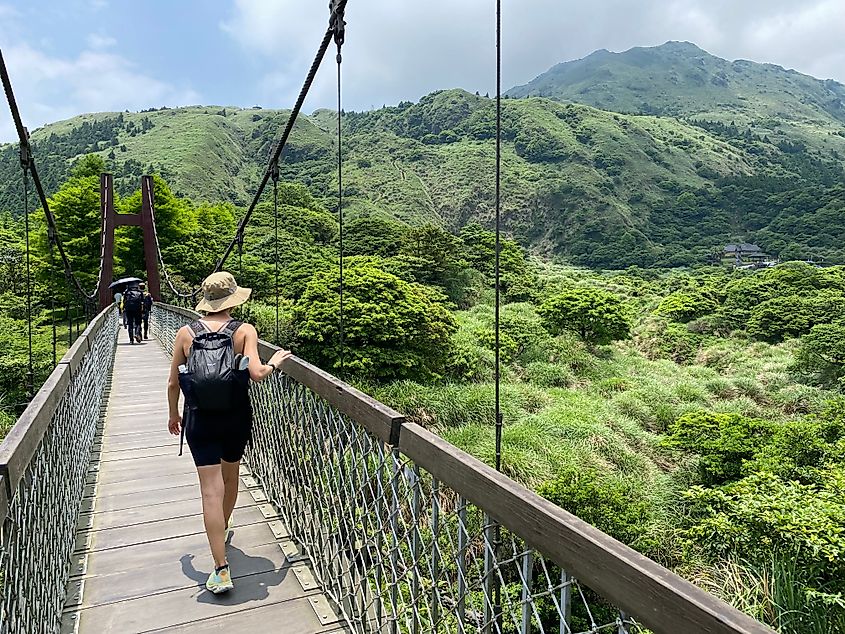 A woman crosses a wooden suspension bridge through a lush mountainscape.