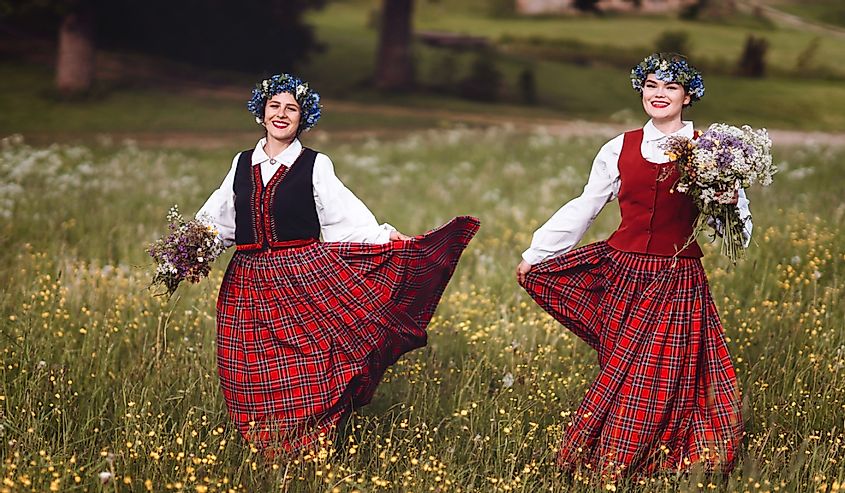 Two Latvian women holding flowers and dressed in traditional clothing in a field of wildflowers