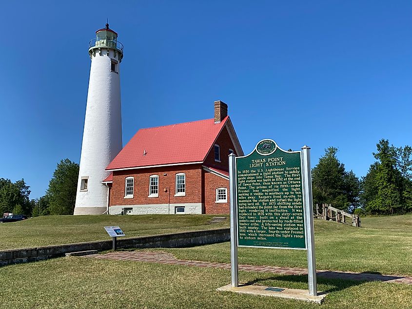 A green historical sign for Tawas Point Light Station stands in front of a red brick dwelling and tall white lighthouse tower