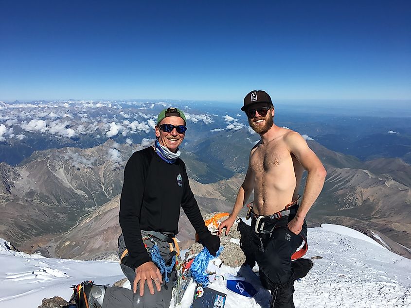 A father and son in climbing gear standing atop the snowy summit of Mount Elbrus in Russia.