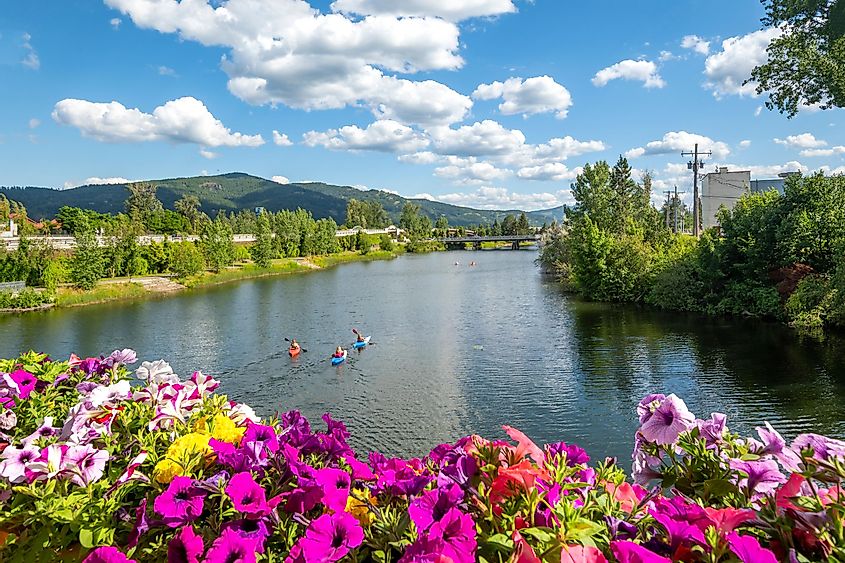Kayakers paddling on Sand Creek River, with the shimmering waters of Lake Pend Oreille in the background, amidst the scenic downtown area of Sandpoint, Idaho on a bright summer day.