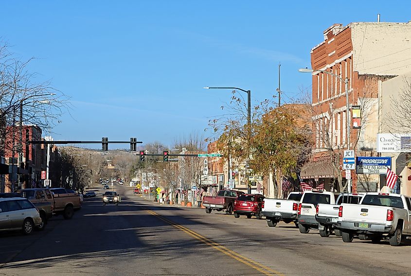 Wide street view of downtown Florence, Colorado, showing historic brick buildings, small shops, parked pickup trucks, traffic lights, and American flags along the sidewalks under a clear blue sky.