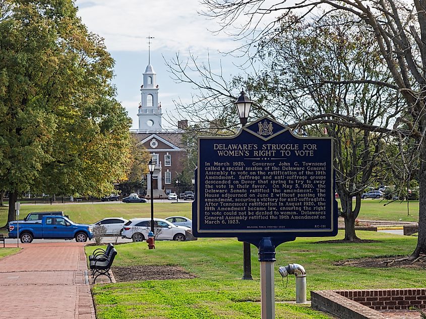 A historical sign about women's voting rights stands on a grassy square in Dover, Delaware, with a colonial-style building and clock tower behind it.