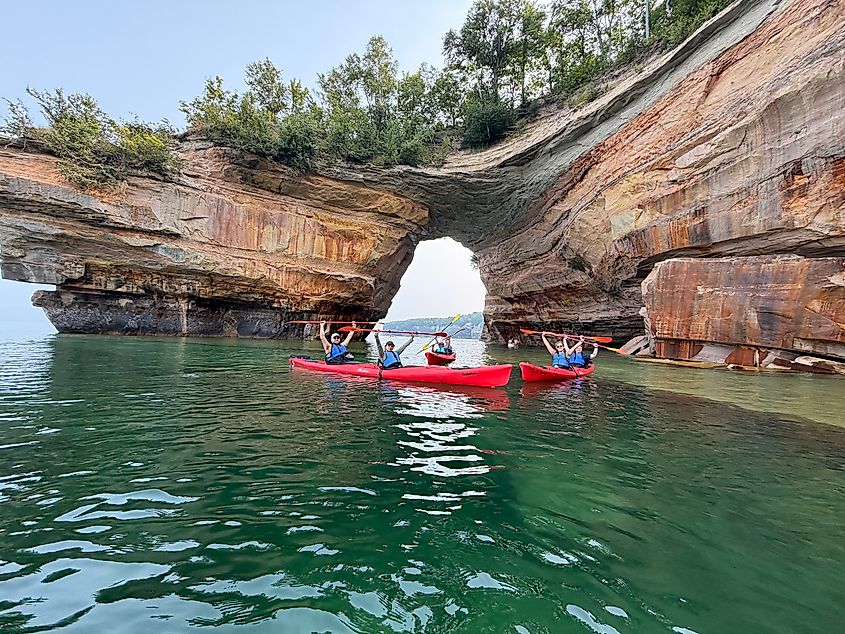 A group of tandem kayakers pose in front of a massive sandstone sea arch. 