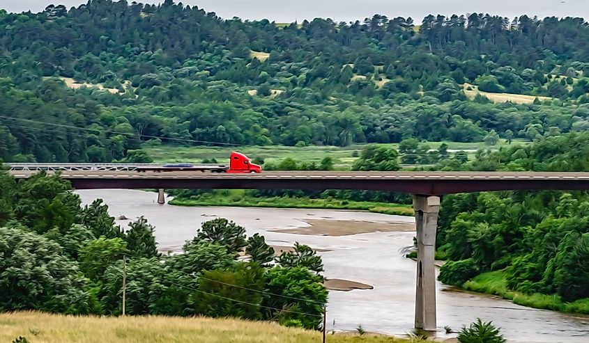 Flatbed truck hauling cargo northwestward along U.S. Route 20 across the Niobrara River toward Valentine, Nebraska, USA, on a summer morning, with digital painting effect