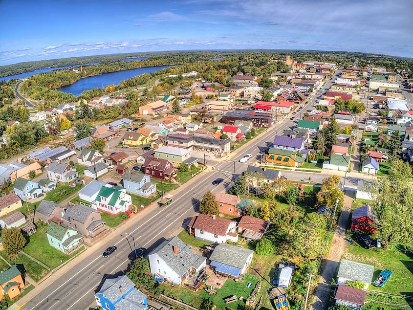 Aerial view of a small town with colorful houses and buildings. A main road cuts through, bordered by lush greenery. A serene lake can be seen in the distance.