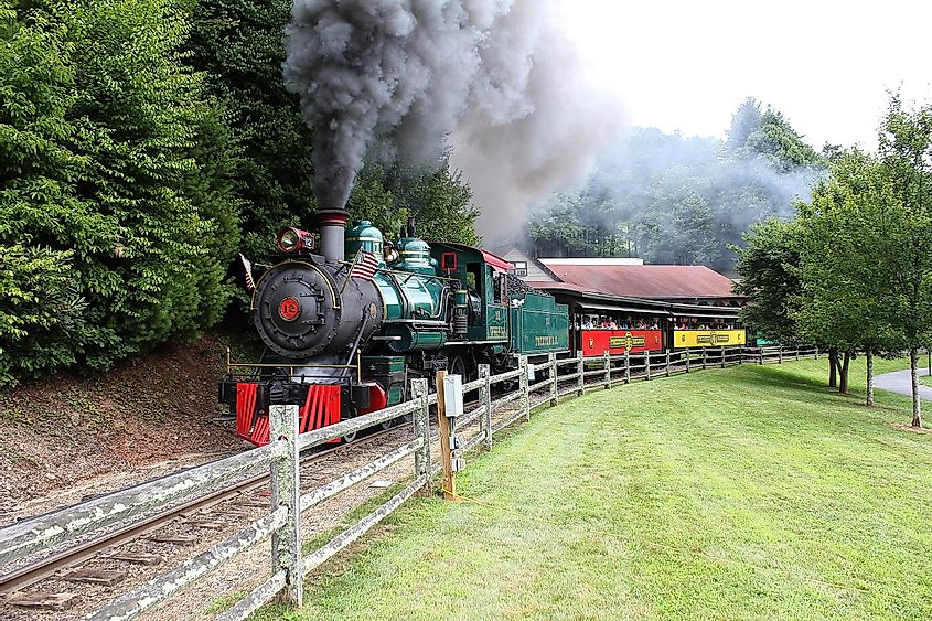Большой игрушечный поезд в парке аттракционов Tweetsie Railroad для детей в горах Смоки с черным дымом и пассажирами-туристами, которые хорошо проводят время.