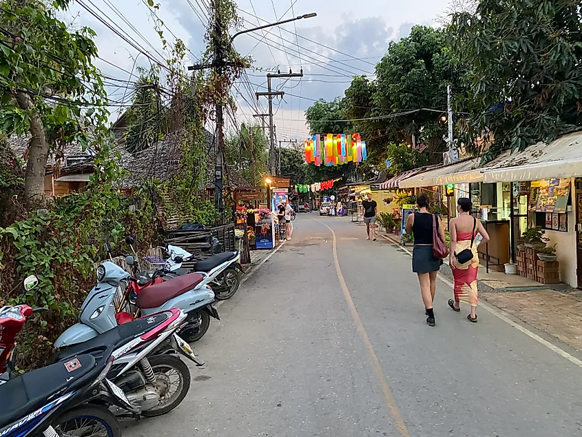 Two women walk down a slim paved road in Asia. Colorful drapings hang from above and motorcycles are parked al along the side.