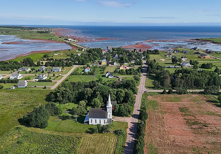 Aerial view of a coastal village with a white church and steeple in the foreground, surrounded by green fields, houses, and a red clay shore by the ocean.