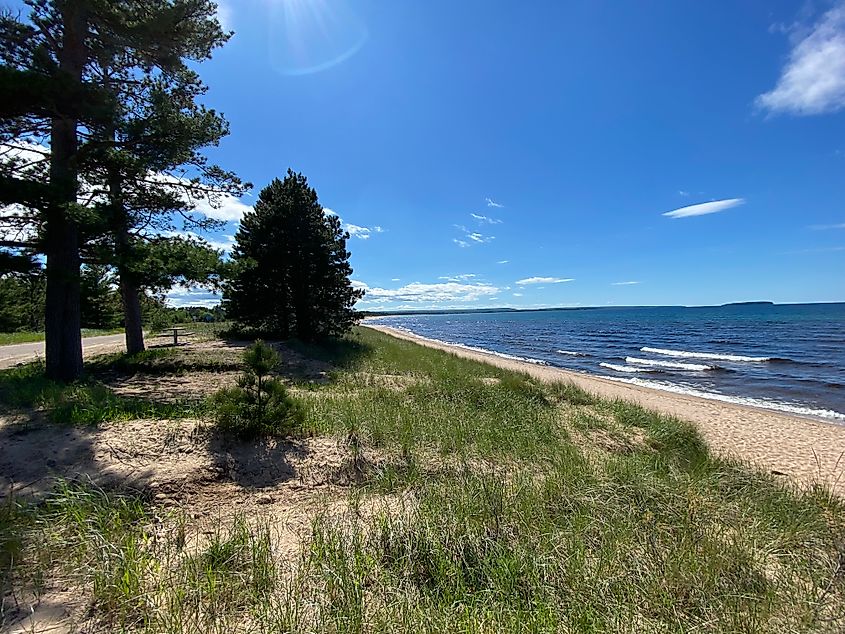 A roadside turnout leading to a long Lake Superior beach, lined with wild dune grass and coniferous trees.