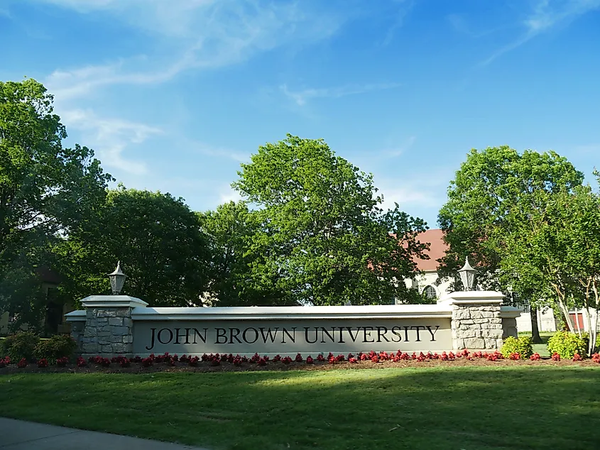 Stone sign reading "John Brown University" surrounded by lush green trees and vibrant flowers under a clear blue sky, conveying a welcoming atmosphere.