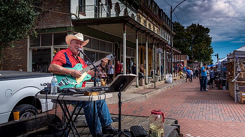 A country folk singer performing on a stage with a guitar on a red brick road during a fall festival in Lebanon, Illinois, USA. Old buildings and craft booths are visible in the background.