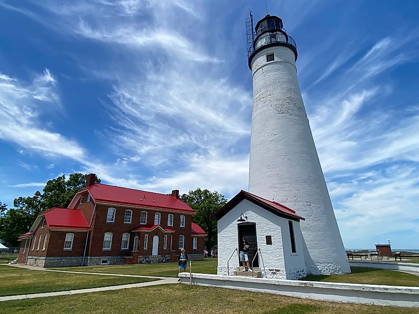 Clouds wisp above a tall white lighthouse next to a large red-brick building.