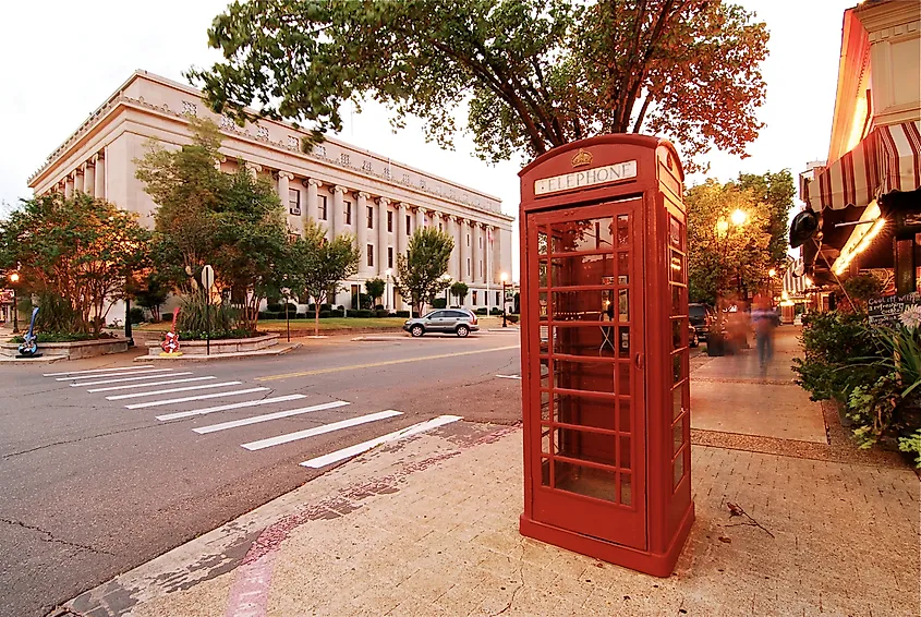 Red phone booth on a sunlit street corner near a large government building. Trees line the street, and warm lights create an inviting, nostalgic atmosphere.