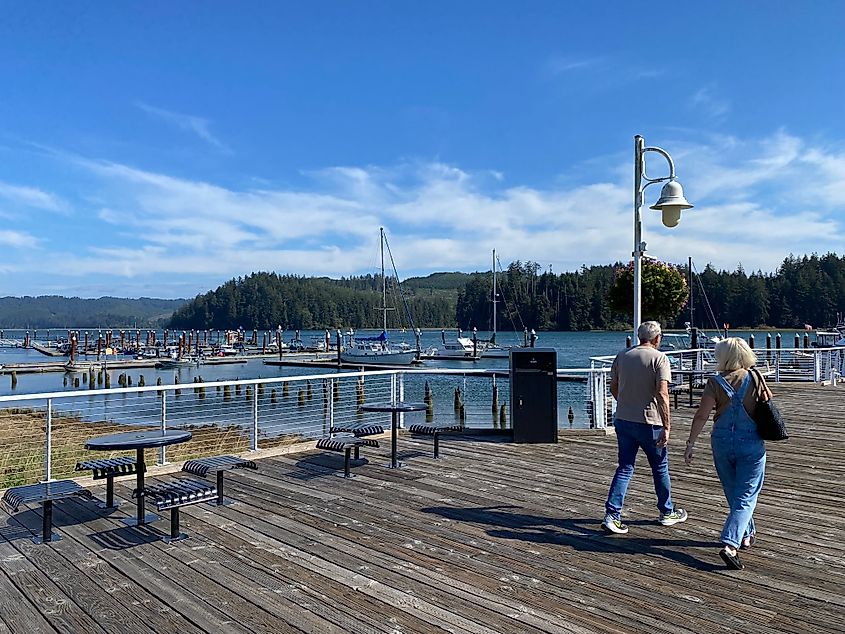 An elderly couple walks along a boardwalk past a small river marina.