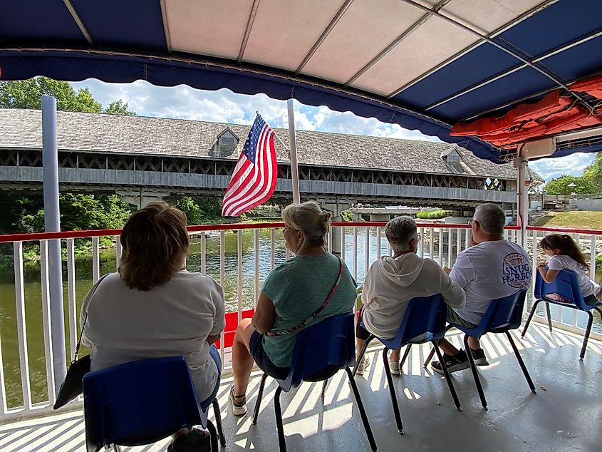 A family looks off the back of a historic riverboat toward a covered wooden bridge that crosses the river