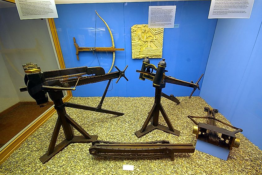 Reconstructions of ancient mechanical artillery displayed at the Saalburg Museum in Hesse, Germany, including a polybolos repeating catapult, bronze-spring and pneumatic catapults, a wooden chain-drive mechanism, and a gastraphetes mounted on the wall