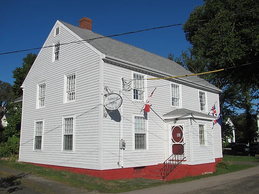Historic two-story white wooden house with a gray roof and red foundation. A sign and flags adorn the exterior. Clear blue sky, inviting atmosphere.